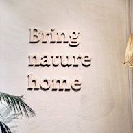 Words in a three-dimensional style on a light-colored wall spell out 'Bring nature home.' A small leafy plant decorates the lower left corner, and a woven wicker lampshade is partially visible on the right.
