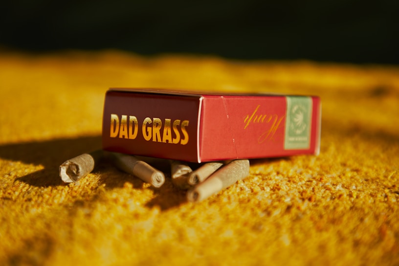 A small red box labeled 'Dad Grass' rests on a textured yellow surface, surrounded by several rolled items resembling cigarettes or joints. The lighting casts a warm, golden hue over the scene, creating distinct shadows.