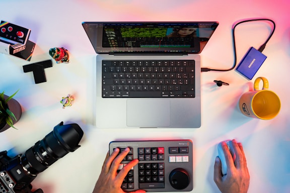 A workspace with a laptop showing video editing software, surrounded by various items including a camera lens, a keyboard with a control panel, an external hard drive, a yellow mug, and a small green plant. Hands are visible operating the keyboard and a mouse.