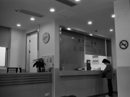 A black-and-white photo of an interior space resembling a hospital or office reception with a counter. A person stands at the counter, possibly filling out paperwork. The counter has a potted plant, hand sanitizer, and signage. A wall clock is mounted above a doorway. Bright lights illuminate the area, and a no-smoking sign is visible.