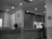 A black-and-white photo of an interior space resembling a hospital or office reception with a counter. A person stands at the counter, possibly filling out paperwork. The counter has a potted plant, hand sanitizer, and signage. A wall clock is mounted above a doorway. Bright lights illuminate the area, and a no-smoking sign is visible.