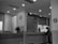 A black-and-white photo of an interior space resembling a hospital or office reception with a counter. A person stands at the counter, possibly filling out paperwork. The counter has a potted plant, hand sanitizer, and signage. A wall clock is mounted above a doorway. Bright lights illuminate the area, and a no-smoking sign is visible.