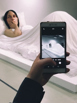 A hyper-realistic sculpture of a person lying in bed, covered with a white sheet. A hand is holding a smartphone, capturing a photo of the scene, with the sculpture displayed on the screen in black and white.