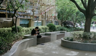 A person is sitting on a curved concrete bench surrounded by greenery. There is a construction site with scaffolding and tarps in the background. Trees and bushes provide a natural canopy and create a tranquil atmosphere.