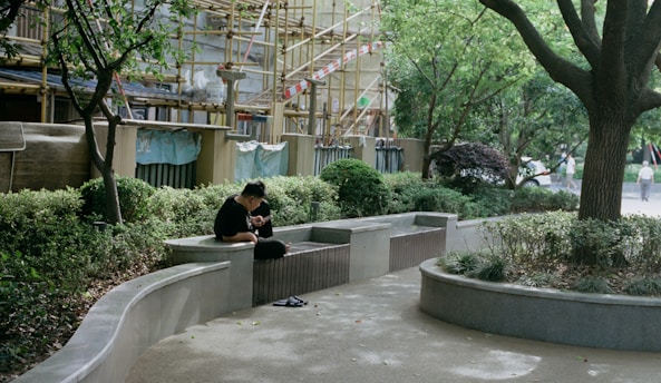A person is sitting on a curved concrete bench surrounded by greenery. There is a construction site with scaffolding and tarps in the background. Trees and bushes provide a natural canopy and create a tranquil atmosphere.