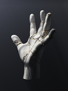 A sculpted hand made of a white material with gold-infused cracks running throughout, seemingly suspended against a dark background. The hand is detailed with realistic textures, and the gold cracks create a striking contrast.