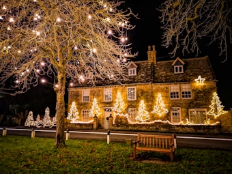 A charming stone house decorated with bright Christmas lights, including star and tree shapes. A large tree in the foreground is adorned with glowing fairy lights, casting a warm and inviting ambiance. A wooden bench sits on a patch of grass, and the scene is set against a dark night sky.