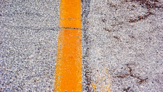 A close-up view of an asphalt road surface featuring a bold yellow line running vertically along the image. The pavement texture is rough, with pebbles and aggregate visible. The line is slightly worn, indicating usage and weather exposure.
