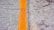 A close-up view of an asphalt road surface featuring a bold yellow line running vertically along the image. The pavement texture is rough, with pebbles and aggregate visible. The line is slightly worn, indicating usage and weather exposure.