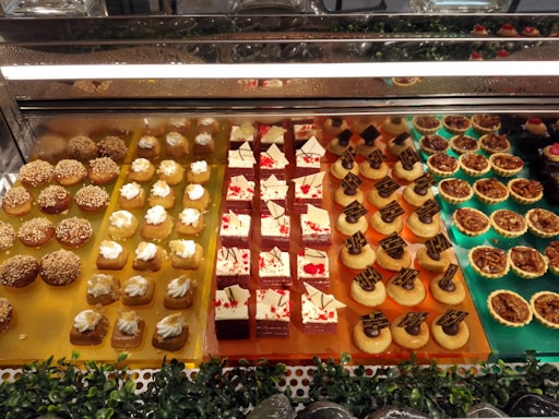An enticing display of assorted desserts is visible, featuring a variety of small pastries and sweets. The arrangement is colorful and meticulously organized in neat rows inside a glass case. Each section boasts different desserts, including small cakes with white frosting, chocolate-topped tarts, nut-covered pastries, and delicate cream-topped squares. The display case is surrounded by small green decorative plants.