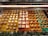 An enticing display of assorted desserts is visible, featuring a variety of small pastries and sweets. The arrangement is colorful and meticulously organized in neat rows inside a glass case. Each section boasts different desserts, including small cakes with white frosting, chocolate-topped tarts, nut-covered pastries, and delicate cream-topped squares. The display case is surrounded by small green decorative plants.