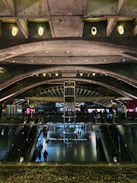 A modern architectural interior featuring a spacious, multi-level atrium with curved concrete arches and a central glass elevator. The space is illuminated with ambient lighting, showcasing escalators and people walking across different levels. Bright digital screens are visible along the walls, adding a vibrant touch to the industrial design.