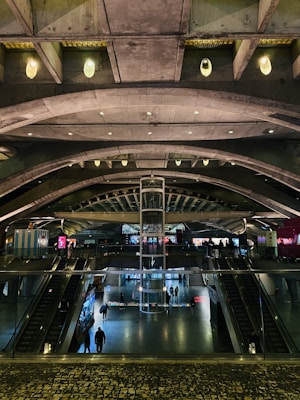 A modern architectural interior featuring a spacious, multi-level atrium with curved concrete arches and a central glass elevator. The space is illuminated with ambient lighting, showcasing escalators and people walking across different levels. Bright digital screens are visible along the walls, adding a vibrant touch to the industrial design.
