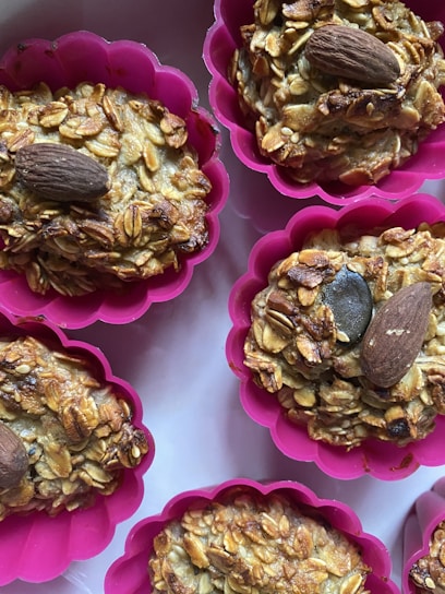 An arrangement of baked muffins topped with almonds, set in bright pink silicone baking cups. The muffins appear to have a grainy texture with visible oats and seeds.