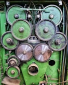 A collection of interconnected metal gears mounted on a green industrial panel. The gears vary in size, with some showing signs of wear and rust. Cables and wires are visible in the background, indicating a mechanical or industrial setup.