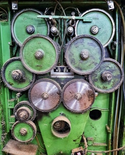 A collection of interconnected metal gears mounted on a green industrial panel. The gears vary in size, with some showing signs of wear and rust. Cables and wires are visible in the background, indicating a mechanical or industrial setup.