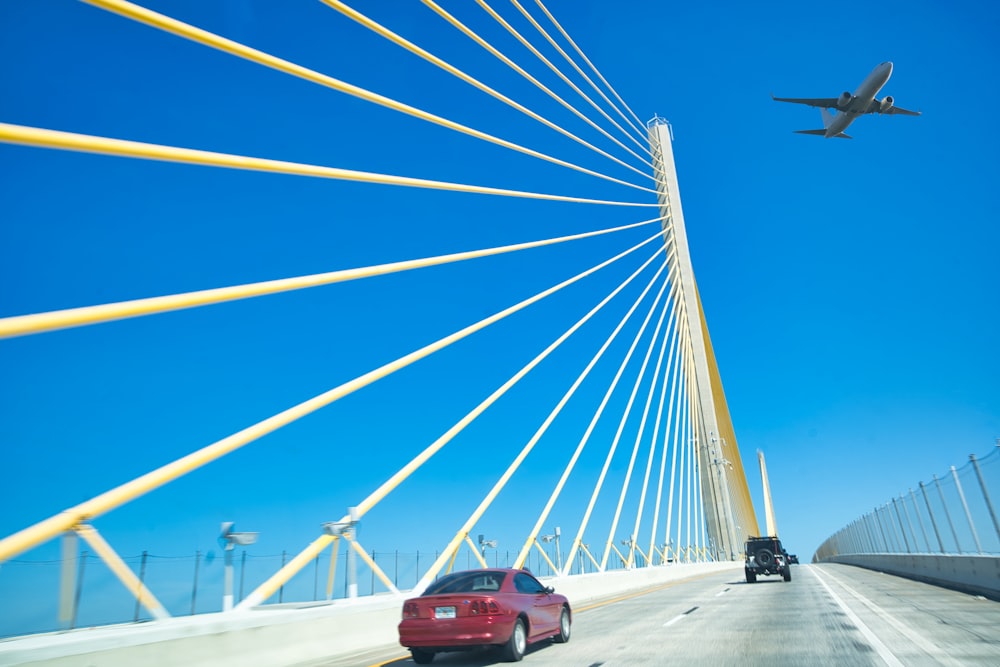 a red car driving across a bridge with a plane flying overhead
