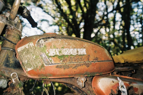 A close-up view of a rusty motorcycle fuel tank with the word 'Suzuki' partially covered in moss and dirt. The tank is orange with signs of wear and age, surrounded by a natural background of blurred trees and foliage.
