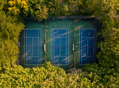 Aerial view of three blue tennis courts surrounded by lush green trees. Players are actively engaged on the courts, and the sunlight casts long shadows, highlighting the surrounding foliage.