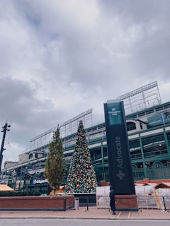 An outdoor scene featuring a large Christmas tree adorned with colorful ornaments. The tree is positioned near a building with visible architectural details, and the sky is overcast. There is a tall sign with the word 'Advocate' on it to the right, and a person dressed in dark clothing is walking nearby.