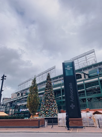 An outdoor scene featuring a large Christmas tree adorned with colorful ornaments. The tree is positioned near a building with visible architectural details, and the sky is overcast. There is a tall sign with the word 'Advocate' on it to the right, and a person dressed in dark clothing is walking nearby.