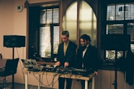 Two individuals standing behind a DJ setup with various audio equipment, including a laptop, mixer, and speakers. The room has a softly lit interior with large windows featuring horizontal blinds, allowing some natural light to filter in. The scene conveys an intimate and focused atmosphere, possibly in a studio or small venue setting.