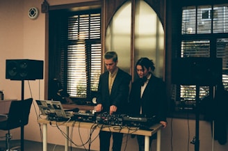 Two individuals standing behind a DJ setup with various audio equipment, including a laptop, mixer, and speakers. The room has a softly lit interior with large windows featuring horizontal blinds, allowing some natural light to filter in. The scene conveys an intimate and focused atmosphere, possibly in a studio or small venue setting.