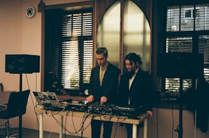 Two individuals standing behind a DJ setup with various audio equipment, including a laptop, mixer, and speakers. The room has a softly lit interior with large windows featuring horizontal blinds, allowing some natural light to filter in. The scene conveys an intimate and focused atmosphere, possibly in a studio or small venue setting.