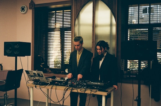 Two individuals standing behind a DJ setup with various audio equipment, including a laptop, mixer, and speakers. The room has a softly lit interior with large windows featuring horizontal blinds, allowing some natural light to filter in. The scene conveys an intimate and focused atmosphere, possibly in a studio or small venue setting.