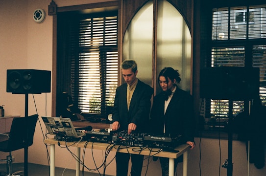 Two individuals standing behind a DJ setup with various audio equipment, including a laptop, mixer, and speakers. The room has a softly lit interior with large windows featuring horizontal blinds, allowing some natural light to filter in. The scene conveys an intimate and focused atmosphere, possibly in a studio or small venue setting.
