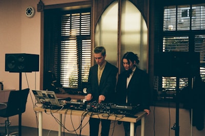 Two individuals standing behind a DJ setup with various audio equipment, including a laptop, mixer, and speakers. The room has a softly lit interior with large windows featuring horizontal blinds, allowing some natural light to filter in. The scene conveys an intimate and focused atmosphere, possibly in a studio or small venue setting.