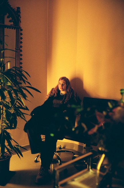 A person is sitting on a black chair in a dimly lit room. The lighting creates a warm and intimate atmosphere with a soft, yellowish hue. There is a large ornate mirror on the left wall and several lush green plants around the room, adding to the cozy and serene setting. The person appears calm and relaxed.