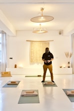 A person wearing a dark hoodie and brown pants is standing in a well-lit, minimalist room with a bright, white interior. The room has yoga mats laid out on the floor, each with a folded blanket and a yoga block. There is a tapestry with a mandala-like pattern hanging on the wall behind the person, flanked by some small decorative objects, including candles, a vase with tall grass-like plants, and a small statue.
