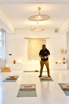 A person wearing a dark hoodie and brown pants is standing in a well-lit, minimalist room with a bright, white interior. The room has yoga mats laid out on the floor, each with a folded blanket and a yoga block. There is a tapestry with a mandala-like pattern hanging on the wall behind the person, flanked by some small decorative objects, including candles, a vase with tall grass-like plants, and a small statue.
