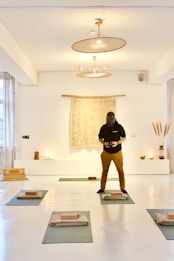 A person wearing a dark hoodie and brown pants is standing in a well-lit, minimalist room with a bright, white interior. The room has yoga mats laid out on the floor, each with a folded blanket and a yoga block. There is a tapestry with a mandala-like pattern hanging on the wall behind the person, flanked by some small decorative objects, including candles, a vase with tall grass-like plants, and a small statue.