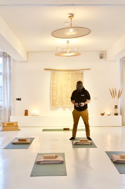 A person wearing a dark hoodie and brown pants is standing in a well-lit, minimalist room with a bright, white interior. The room has yoga mats laid out on the floor, each with a folded blanket and a yoga block. There is a tapestry with a mandala-like pattern hanging on the wall behind the person, flanked by some small decorative objects, including candles, a vase with tall grass-like plants, and a small statue.