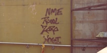 Graffiti is spray-painted on an industrial-looking wall with words in a bold and uneven style. The wall is aged with spots and marks. There is a metal railing running horizontally below the graffiti, and a scaffold or rack is visible on the right side. A plastic bag or tarp is also partially visible on the right.
