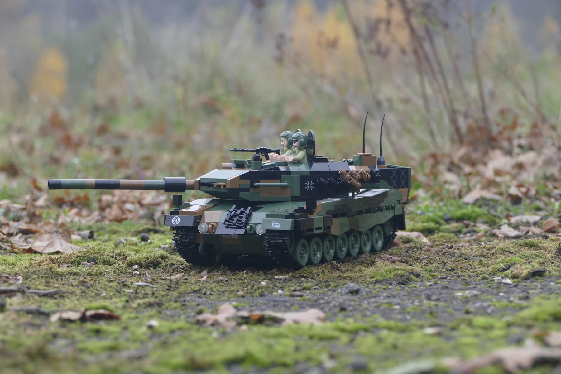 A green and black camouflaged model battle tank on moss and fallen leaves, its gun pointing left