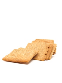 Several square, lightly textured crackers are arranged in a staggered stack on a white background. The crackers have a golden-brown color with a slightly rough surface.
