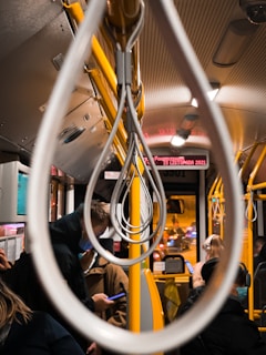 Interior of a bus with yellow handrails and straps, occupied by several passengers. Some individuals are standing, while others are seated. The focus is on the hand straps creating a symmetrical pattern. Outside the window, city lights and traffic blur by, indicating movement during nighttime.