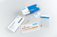A collection of items related to a test kit, including a white and blue box labeled 'testalize.me', a sealed return envelope, a small vial, an information booklet, a code card, a pipette, and a clear plastic bag with a blue strip.