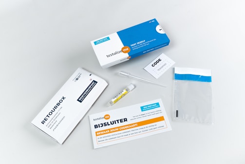A collection of items related to a test kit, including a white and blue box labeled 'testalize.me', a sealed return envelope, a small vial, an information booklet, a code card, a pipette, and a clear plastic bag with a blue strip.