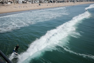A surfer rides a wave near the shore of a busy beach lined with buildings. The water is a deep blue-green, topped with white frothy waves. People are scattered along the sandy beach, some under umbrellas, while others wade in the shallow waters.