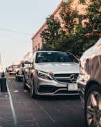 A row of luxury cars, including a silver Mercedes-Benz, is parked on a cobblestone street. Trees and buildings line the street, and there are people visible inside the front car. The lighting suggests early evening with a warm, soft glow.