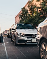 A row of luxury cars, including a silver Mercedes-Benz, is parked on a cobblestone street. Trees and buildings line the street, and there are people visible inside the front car. The lighting suggests early evening with a warm, soft glow.