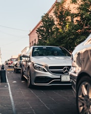 A row of luxury cars, including a silver Mercedes-Benz, is parked on a cobblestone street. Trees and buildings line the street, and there are people visible inside the front car. The lighting suggests early evening with a warm, soft glow.