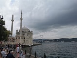 A scenic waterfront view showcases a mosque with two minarets in the foreground, adjacent to a body of water. People gather along the water's edge, engaging in leisure activities. In the background, a large suspension bridge stretches across the scene under a cloudy sky, with hills visible on the horizon.
