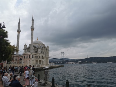 A scenic waterfront view showcases a mosque with two minarets in the foreground, adjacent to a body of water. People gather along the water's edge, engaging in leisure activities. In the background, a large suspension bridge stretches across the scene under a cloudy sky, with hills visible on the horizon.
