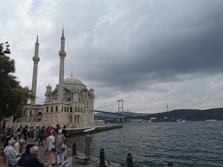 A scenic waterfront view showcases a mosque with two minarets in the foreground, adjacent to a body of water. People gather along the water's edge, engaging in leisure activities. In the background, a large suspension bridge stretches across the scene under a cloudy sky, with hills visible on the horizon.