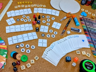 A collection of board game components and crafting materials are spread out on a cork board surface. There are playing cards, dice in various colors and sizes, wooden letter tiles spelling 'BOARDGAMES', 'DRAW', and 'WIN', colored buttons, a variety of markers and colored pencils, scissors, glue stick, and circular cardboard cutouts. The setting is vibrant and full of an assortment of gaming and crafting items.